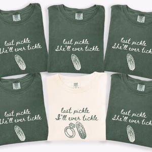 May include: A collection of six t-shirts, five in a muted green and one in cream. Each shirt features the phrase "last pickle she'll ever tickle" with a pickle illustration, except for one cream shirt that says "last pickle I'll ever tickle" with a ring and pickle.