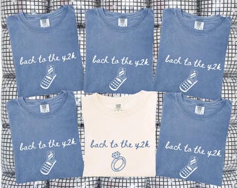 May include: Six t-shirts, five in faded blue and one in cream, are displayed. Each shirt features the text "bach to the y2k" with a flip phone graphic, except for one cream shirt with a diamond ring graphic.