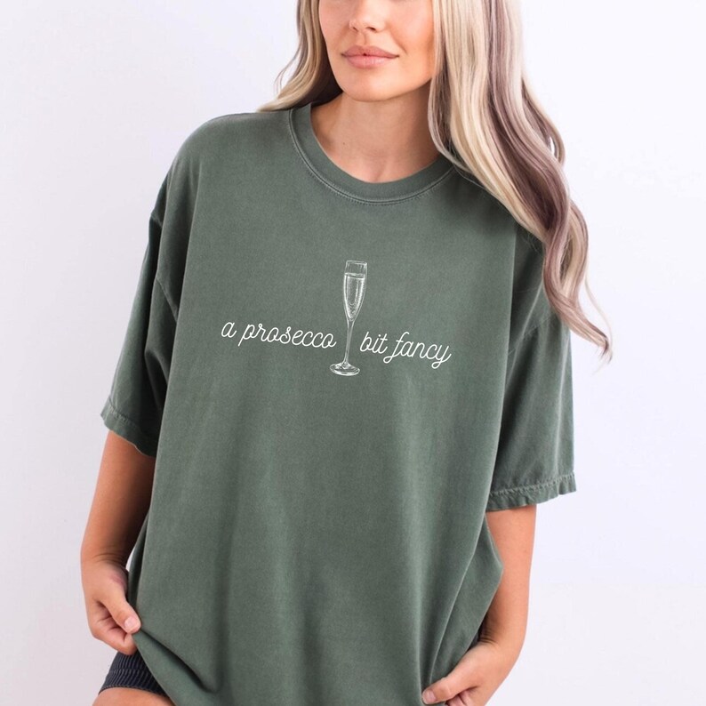 May include: A green t-shirt with a white graphic of a champagne flute and the text "a prosecco bit fancy".