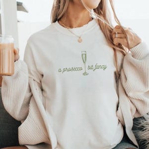 May include: White sweatshirt with a green illustration of a champagne flute and the text "a prosecco bit fancy".
