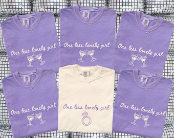 May include: Six t-shirts, five violet and one ivory, with the text "One less lonely girl" and a cocktail glass graphic. The ivory shirt features a ring graphic. Each shirt has a necklace. The shirts are displayed on a reflective surface.