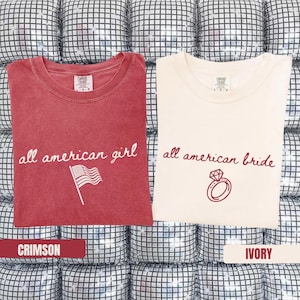 May include: Two t-shirts, one crimson and one ivory, with the words "all american girl" and "all american bride" printed on them. The crimson shirt has an American flag graphic, and the ivory shirt has a ring graphic.