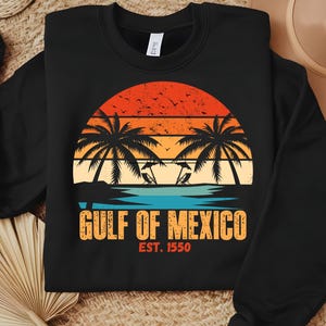 Könnte beinhalten: Schwarzer Pullover mit einem Vintage-Grafikdruck von Palmen und einem Sonnenuntergang über dem Meer. Der Text "Golf von Mexiko Est. 1550" ist unter dem Grafikdruck aufgedruckt.