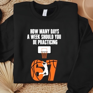 Peut inclure: Sweat-shirt noir avec le texte "HOW MANY DAYS A WEEK SHOULD YOU BE PRACTICING" au-dessus d'un panier de basket et le chiffre "67" en orange avec un motif de ballon de basket. Silhouette d'un joueur de basket-ball en train de dunker.