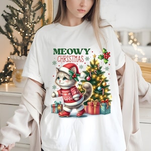 May include: White t-shirt with a Christmas cat design. The cat is wearing a Santa hat and holding a gift. The text "MEOWY CHRISTMAS" is printed on the shirt.