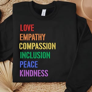May include: Black crewneck sweatshirt with the words "LOVE, EMPATHY, COMPASSION, INCLUSION, PEACE, KINDNESS" in rainbow colors. The text is in a stencil font. The sweatshirt is folded on a woven surface.