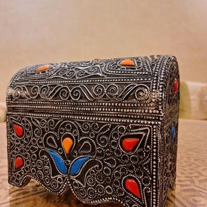 May include: A black wooden box with intricate silver filigree designs and colorful gemstones. The box has a rounded top and a scalloped base.