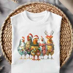May include: White t-shirt featuring a group of five chickens dressed for Christmas. The chickens are adorned with Santa hats, reindeer antlers, and festive lights. The chickens are wearing scarves and have colorful lights wrapped around them.