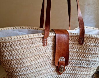 May include: A large, woven straw tote bag with brown leather straps and a buckle closure. The bag has a white lining and is perfect for carrying everyday essentials.