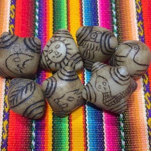 Puede incluir: Siete pequeñas cuentas de cerámica triangulares de color beige con varios diseños. Los diseños incluyen un sol, un pájaro, una serpiente, una hoja, una luna y un patrón geométrico. Las cuentas están dispuestas sobre una tela de rayas de colores.