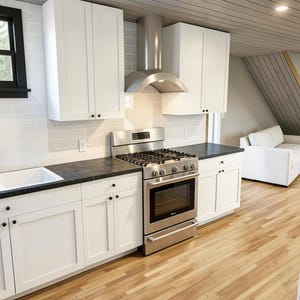 May include: A modern kitchen with white cabinets, a stainless steel stove and range hood, and a black countertop. The kitchen has a white sink and a window with a black frame. The flooring is light-colored wood.