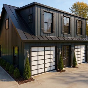 May include: A two-story, dark gray building with a black roof and white garage doors. The building has multiple windows and is surrounded by trees with yellow leaves. The driveway is concrete.