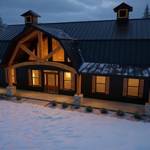 May include: A dark-sided house with a black metal roof, two cupolas, and a large timber-framed front porch entrance. The exterior features vertical siding, stone-based support columns, and warm interior lighting visible through the windows.
