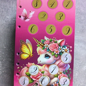 Könnte beinhalten: Rosa Notizbuch mit einer Einhorn-Illustration und Schmetterlingen. Das Einhorn hat einen Blumenkranz und ist von Rosen umgeben. Goldene Kreise mit den Zahlen 1-9 sind oben angeordnet. Das Notizbuch hat eine Spiralbindung auf der linken Seite.