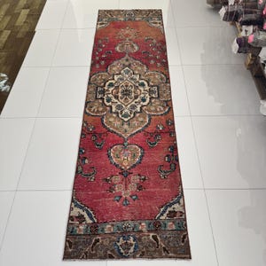 May include: A long, rectangular runner rug featuring a vibrant red base color with intricate floral and geometric patterns in shades of beige, dark brown, and teal. The design includes a central medallion and decorative borders at both ends.