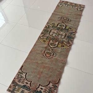 Könnte beinhalten: Ein rechteckiger, abgenutzt aussehender Teppich mit einem zentralen Blumenmuster in Grün-, Rosa- und Beigetönen. Der Teppich hat ein verblasstes Aussehen mit einem Rand mit einem Muscheldesign. Der Teppich liegt auf einem weißen Fliesenboden.