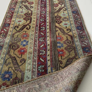 May include: A rectangular runner rug featuring a complex pattern of geometric shapes and floral motifs in shades of olive green, deep red, and blue. The rug has a thick, dark brown central vertical band and a visible woven backing at the corner.