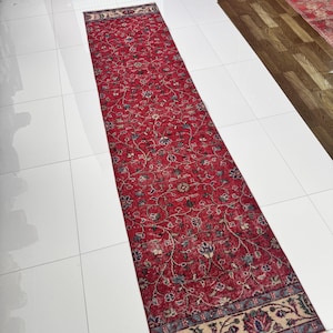 Może przedstawiać: Długi, prostokątny czerwony dywan ze skomplikowanym wzorem kwiatowym. Dywan ma centralne pole w kolorze czerwonym ze szczegółowymi wzorami kwiatowymi w odcieniach niebieskiego, zielonego i beżowego. Obramowanie ma kontrastowy wzór. Dywan leży na białej podłodze z płytek.
