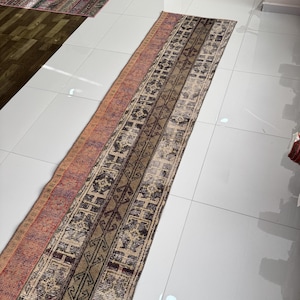 Pode incluir: Um tapete longo e retangular com uma aparência desbotada e vintage. O tapete apresenta um design central com padrões geométricos em tons de castanho, bege e creme, ladeado por uma secção de tons avermelhados-alaranjados e roxos. O tapete está colocado sobre um chão de azulejos brancos.