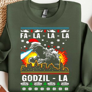 Può includere: Felpa verde scuro con una grafica di Godzilla con un cappello da Babbo Natale, che sputa fiamme blu, e il testo "FA-LA-LA-LA" e "GODZIL-LA". Il design include fiocchi di neve, alberi di Natale e carri armati.
