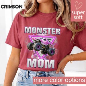 May include: Crimson red t-shirt with the words "MONSTER MOM" in silver block letters. A pink monster truck with a shark mouth design is centered on a pink triangle. The words "super soft" and "more color options" are also visible.
