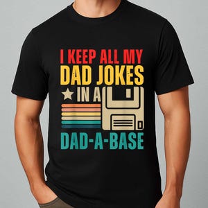Op de afbeelding: Zwart t-shirt met de tekst "I KEEP ALL MY DAD JOKES IN A DAD-A-BASE" in rood, geel en blauwgroen. Het ontwerp bevat een ster, horizontale strepen en een floppy disk-afbeelding.