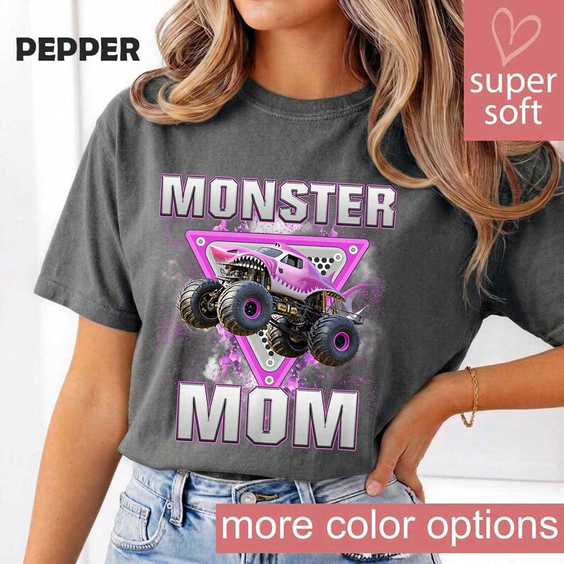 May include: Dark gray t-shirt with the words "MONSTER MOM" in white and pink lettering. A pink monster truck graphic is centered on the shirt. The word "PEPPER" is in the upper left corner. The words "more color options" are at the bottom.