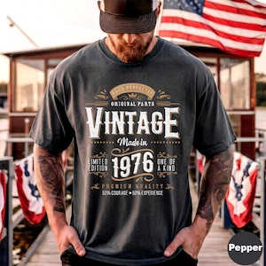 May include: Dark gray t-shirt with the text "VINTAGE Made in 1976." The shirt has a distressed look with a vintage design. The text includes "Original Parts," "Limited Edition," and "One of a Kind." The shirt is worn by a person.