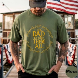 Puede incluir: Camiseta verde musgo con el texto dorado "ONE DAD TO RULE THEM ALL" dentro de un diseño circular dorado. La camiseta tiene mangas cortas y cuello redondo. La palabra "Moss" está impresa en una fuente pequeña y oscura en la parte inferior derecha.