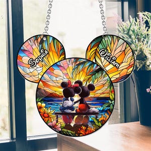 Puede incluir: Un atrapasol de vidrio emplomado con Mickey y Minnie Mouse en una escena de puesta de sol con palmeras. El atrapasol está hecho de tres piezas redondas de vidrio con los nombres "Sophia" y "William" en dos de las piezas.