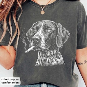 Può includere: Una t-shirt a maniche corte color grigio antracite con un'illustrazione a linee bianche di un cane con orecchie pendenti che tiene una sigaretta in bocca. La grafica è centrata sul petto del capo, caratterizzato da un girocollo.
