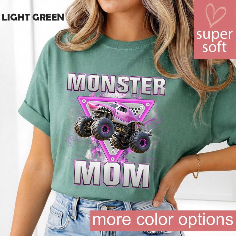 May include: Light green t-shirt with the words "MONSTER MOM" in white and pink. The shirt features a pink monster truck graphic inside a pink triangle. The words "super soft" and "more color options" are also visible.