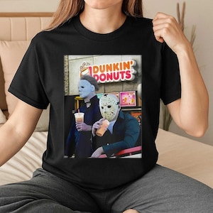 May include: Black t-shirt with a graphic of two horror film characters, one in a white mask and the other a pale mask, holding iced coffees in front of a Dunkin' Donuts sign.