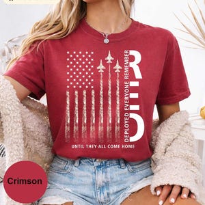 Può includere: T-shirt color cremisi con un design a bandiera americana invecchiata e jet in ascesa. La maglietta include il testo "REMEMBER" in verticale, e "DEPLOYED EVERYONE" e "UNTIL THEY ALL COME HOME".