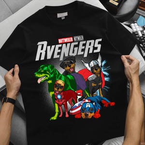 May include: Black t-shirt featuring a graphic of Rottweiler dogs dressed as Avengers characters. The word "Avengers" is in large white letters, with "Rottweiler" above. The dogs are in various superhero costumes, including Hulk, Iron Man, Thor, and Captain America.