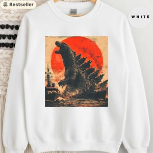 Può includere: Felpa bianca con grafica di Godzilla che ruggisce davanti a un sole rosso, con navi da guerra nell'oceano. Il design è di ispirazione vintage, con un aspetto invecchiato.