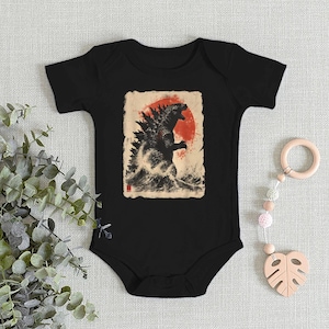 May include: Black baby bodysuit featuring a Godzilla graphic. The design includes a red sun and a black and white illustration of Godzilla emerging from the water. The bodysuit is made of soft fabric and has short sleeves.