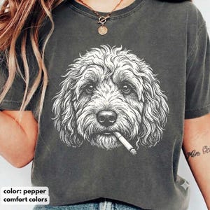 May include: A charcoal grey short-sleeved t-shirt featuring a white line-art illustration of a curly-haired dog with a cigarette in its mouth. The shirt has a crew neckline and a vintage-style washed finish, showcasing a detailed canine portrait.