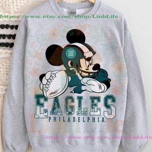 Könnte beinhalten: Ein hellgraues Sweatshirt mit einer Cartoonfigur in Football-Uniform, die einen Football hält. Das Wort "EAGLES" ist in großen, teal-farbenen Buchstaben gedruckt, darunter "PHILADELPHIA". Das Design enthält einen Platz für ein Logo.