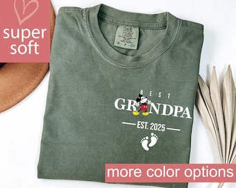 May include: Olive green t-shirt with the words "BEST GRANDPA" in white, featuring a Mickey Mouse graphic. Below the text is "EST. 2025" and two white footprints. The shirt has a crew neck and a tag that says "COMFORT COLORS".