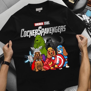 Puede incluir: Camiseta negra con un diseño de dibujos animados de Cocker Spaniels disfrazados de superhéroes. El texto "COCKER SPANIEL" está encima de la palabra "COCKERSPAVENGERS" en una fuente estilizada. El diseño incluye perros con disfraces.