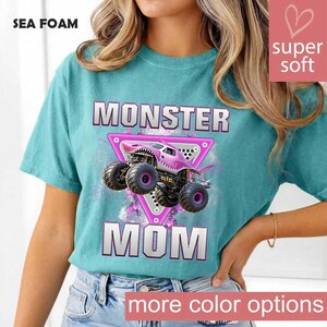 May include: Sea foam green t-shirt with the words "MONSTER MOM" in silver and pink. A pink monster truck graphic is centered within a pink triangle. The shirt has a "super soft" label and a "more color options" label.