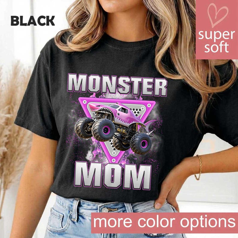 May include: Black t-shirt with a graphic of a pink monster truck with the words "MONSTER MOM" in white block letters. The design includes a pink triangle and the text "super soft" in a pink box. The words "more color options" are at the bottom.