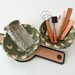 May include: A clear glass jar and baking tools rest on a green fabric with a white chicken pattern. The tools include a whisk, spatula, and wooden utensils. The items are on a wooden cutting board.