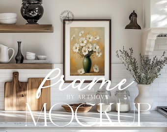May include: A white framed picture of a vase with white flowers in a kitchen setting. The picture is hanging on a white tiled wall above a white countertop with a wooden cutting board, a bowl of nuts, and two glass jars.