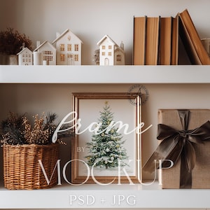 Puede incluir: Una impresión enmarcada en oro de un árbol de Navidad con una estrella, exhibida en un estante blanco. Una cesta tejida con ramas decorativas, un regalo envuelto con una cinta marrón y casas blancas en miniatura con ventanas iluminadas también están en el estante.