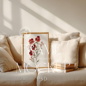 May include: A framed watercolour painting of red flowers with green stems and leaves on a white background. The frame is a light wood colour and is sitting on a white sofa with a stack of books in front of it.