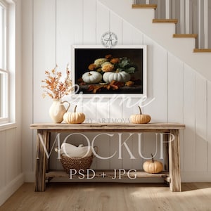 Puede incluir: Una consola rústica de madera exhibe una pintura enmarcada de naturaleza muerta de calabazas y flores. Pequeñas calabazas y un jarrón con follaje seco están dispuestos sobre la mesa. Una cesta tejida con un paño blanco está en el estante inferior. Tema de cosecha de otoño.