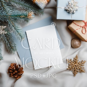 May include: A blank white card and light blue envelope with the text "CARD MOCKUP" are displayed on a soft, white fabric. The scene includes a pine cone, gold snowflake, and wrapped gifts, suggesting a holiday theme.