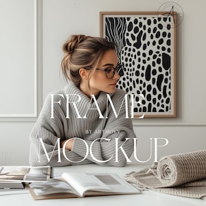 Puede incluir: Una mujer con gafas y un suéter gris está sentada en un escritorio blanco con un libro abierto y una manta enrollada. Una obra de arte abstracta en blanco y negro enmarcada cuelga de la pared. Se muestra el texto "FRAME MOCKUP".